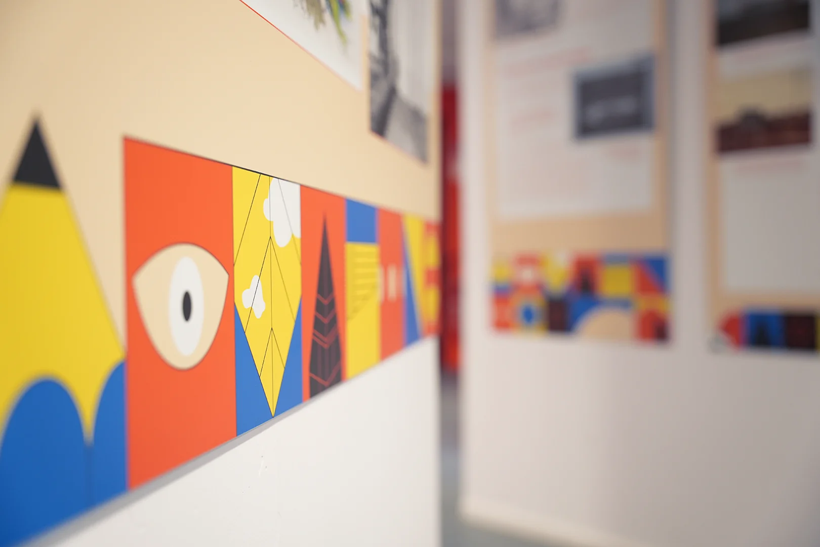 Détail du bas d'un panneau installé dans l'exposition. Focus sur la frise multicolore qui habille le bas des panneaux : elle est composée de blocs carrés comportant des illustrations dessinées au trait noir (crayon, fenêtre, détail d'architectures, formes géométriques). À l'arrière-plan, flou, on devine deux autres panneaux.